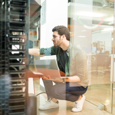 Network administrator with notebook computer sitting in data center room and working with networking device on rack cabinet