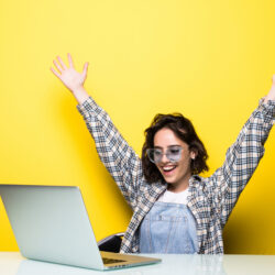 Cheerful woman in glasses holding laptop and showing fist up excited with win on yellow background.