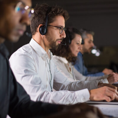 Side view of confident call center operator talking with client. Caucasian young man in eyeglasses typing on laptop while serving client. Call center concept