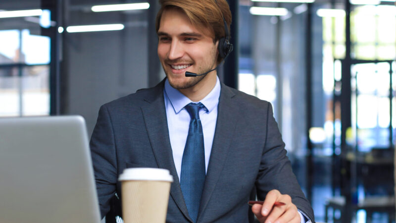 Smiling male call-center operator with headphones sitting at modern office, consulting online information in a laptop, looking up information in a file in order to be of assistance to the client