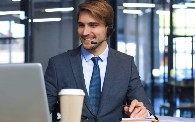 Smiling male call-center operator with headphones sitting at modern office, consulting online information in a laptop, looking up information in a file in order to be of assistance to the client