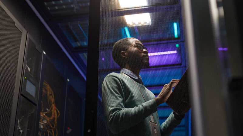 Low angle portrait of African American data engineer working with supercomputer in server room lit by blue light, copy space