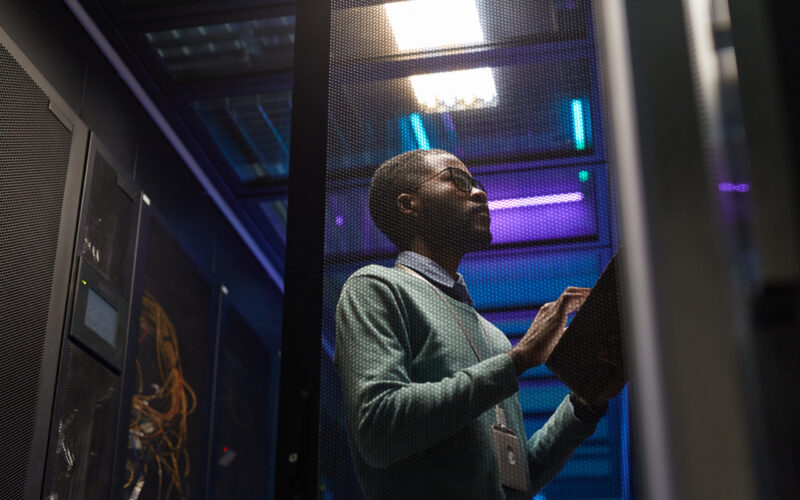 Low angle portrait of African American data engineer working with supercomputer in server room lit by blue light, copy space