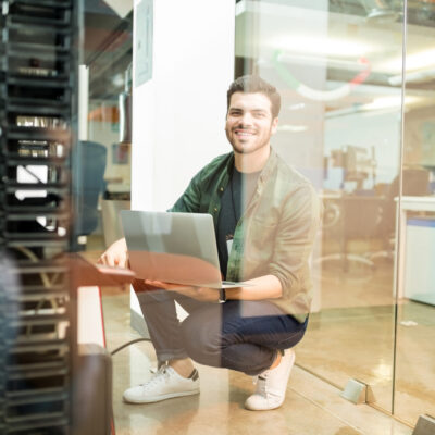 Portrait of happy young male network engineer with laptop in hand working in datacenter.